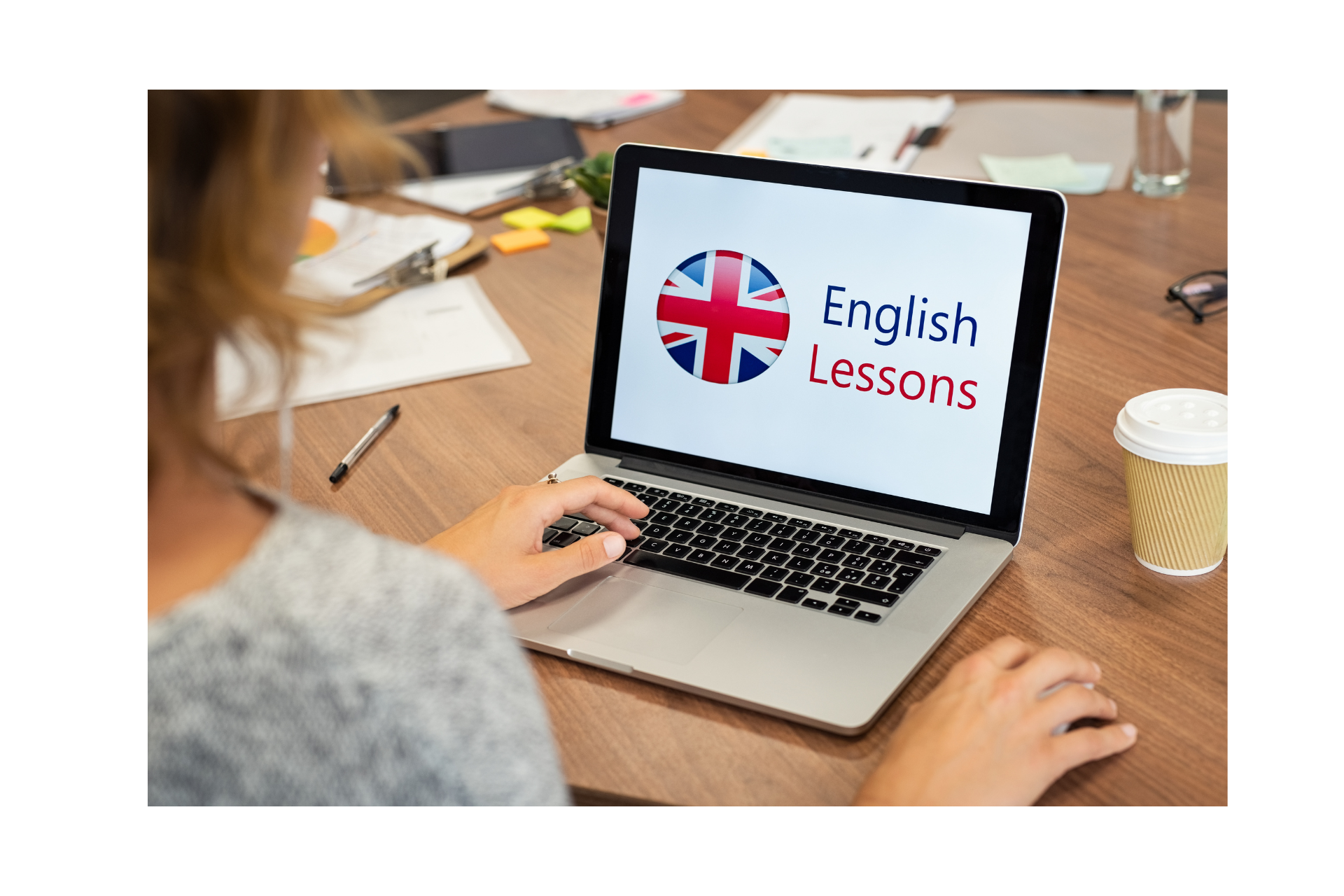 Na zdjęciu laptop z wyświetlonymi na ekranie angielskimi napisami oraz flagą brytyjską. Obok laptopa leżą przedmioty biurowe. Po lewej stronie głowa i ramię kobiety używającej laptopa.