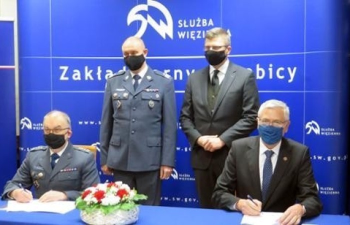 Czwórka mężczyzn w maseczkach przy stole. Dwóch siedzi i podpisuje dokumenty. W tle logotyp Służby Więziennej.