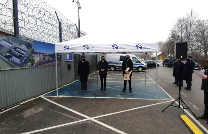  Three men with their face masks at the microphones under the tent. Barbed-wired fence on the left and uniformed people on the right. 