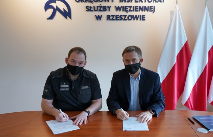 Two men with their face masks on signing a contract. One of the is wearing his uniform, the other one a jacket. A Polish flag and the Prison Service logo behind them. 