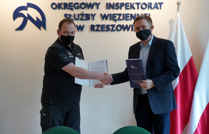 Two men with their face masks on shaking their hands. Polish flags and the Prison Service logo behind them. 