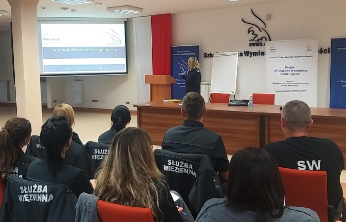 Grupa studentów siedzi na krzesłach i słucha prowadzącej podczas wykładu. Po lewej stronie ekran z wyświetlanym obrazem. W tle polska flaga i logotypy Szkoły Wyższej Wymiaru Sprawiedliwości. Grupa studentów siedzi na krzesłach i słucha prowadzącej podczas wykładu. Po lewej stronie ekran z wyświetlanym obrazem. W tle polska flaga i logotypy Szkoły Wyższej Wymiaru Sprawiedliwości.