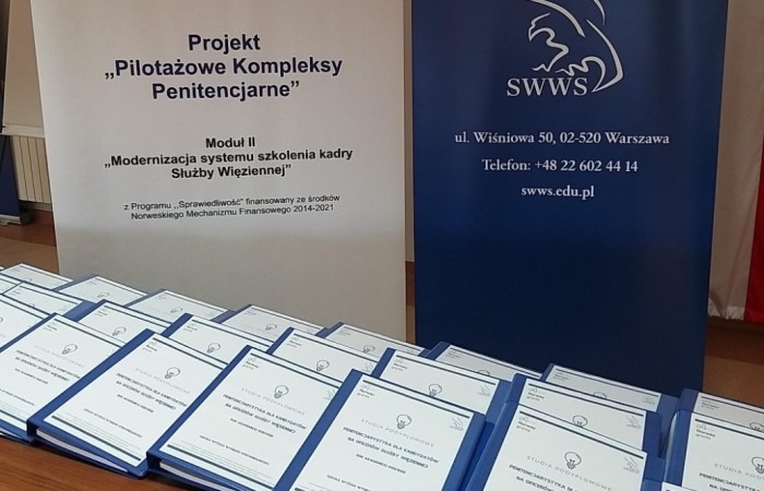 Na stole o brązowym blacie leżą ułożone w rzędach segregatory z białymi okładkami. Za stołem banery z logotypami Szkoły Wyższej Wymiaru Sprawiedliwości. Na stole o brązowym blacie leżą ułożone w rzędach segregatory z białymi okładkami. Za stołem banery z logotypami Szkoły Wyższej Wymiaru Sprawiedliwości.