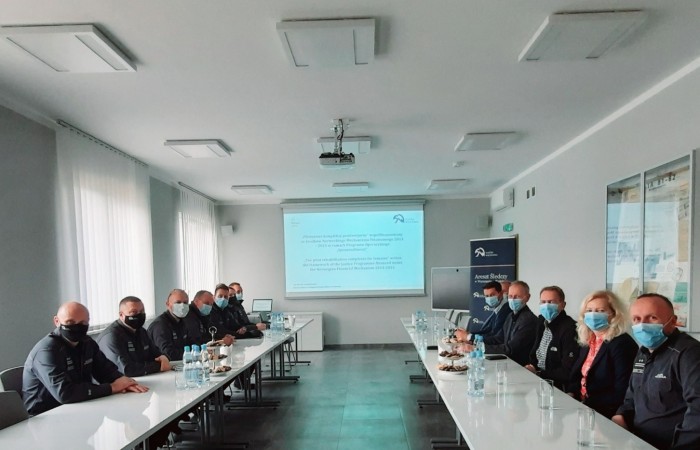 Grupa ludzi w maseczkach siedzi przy stołach po dwóch stronach sali. W tle prezentacja multimedialna z logotypem Służby Więziennej. Grupa ludzi w maseczkach siedzi przy stołach po dwóch stronach sali. W tle prezentacja multimedialna z logotypem Służby Więziennej.