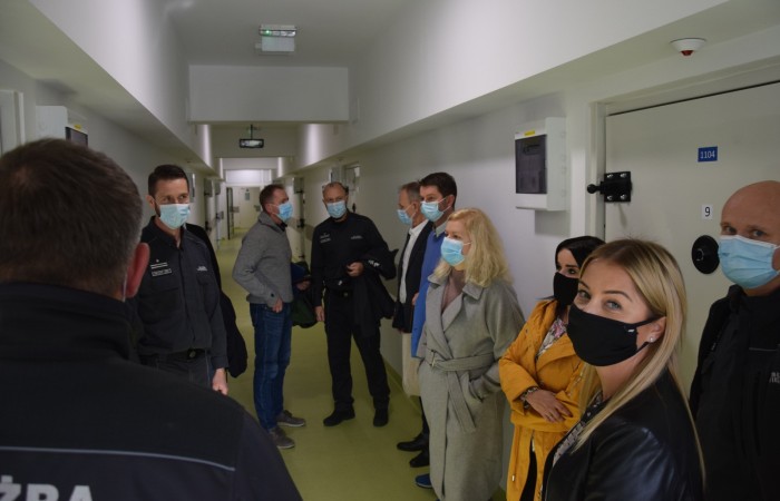 Grupa ludzi w maseczkach rozmawia ze sobą wewnątrz zakładu karnego. Parę osób w mundurach Służby Więziennej. Po prawej i lewej stronie wejścia do cel. Grupa ludzi w maseczkach rozmawia ze sobą wewnątrz zakładu karnego. Parę osób w mundurach Służby Więziennej. Po prawej i lewej stronie wejścia do cel.