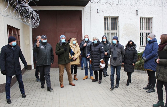 In the photo, a group of people wearing face masks, dressed in jackets, are standing in front of the correctional facility with barbed wire and barred windows. In the photo, a group of people wearing face masks, dressed in jackets, are standing in front of the correctional facility with barbed wire and barred windows.