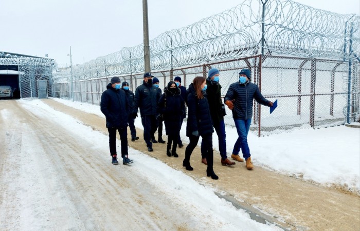 A group of people wearing face masks, dressed in jackets, is talking outside, in the snowy scenery. Behind them, there is a wall and a barbed wire fence. A group of people wearing face masks, dressed in jackets, is talking outside, in the snowy scenery. Behind them, there is a wall and a barbed wire fence.