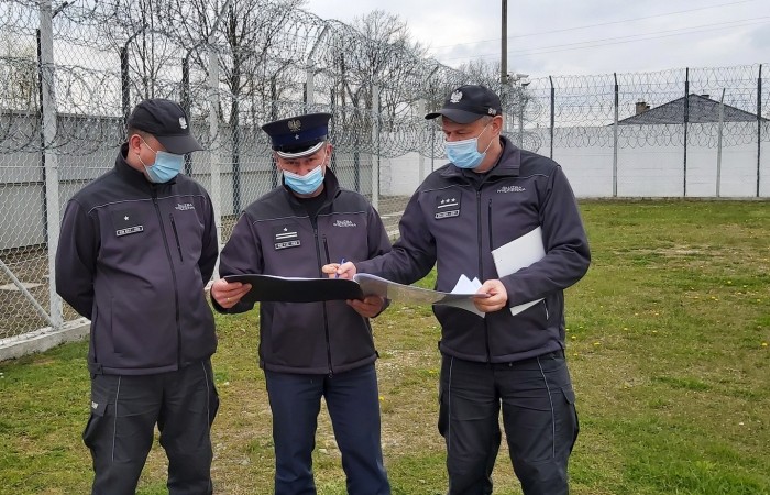 Trzech funkcjonariuszy w maseczkach stoi na trawie. Przeglądają dokumenty. W tle siatka i drut kolczasty. Trzech funkcjonariuszy w maseczkach stoi na trawie. Przeglądają dokumenty. W tle siatka i drut kolczasty.