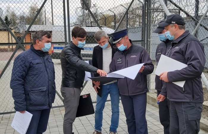 Grupa sześciu mężczyzn w maseczkach spogląda na dokument. Trzech w umundurowaniu. Za nimi wjazd do zakładu karnego. Grupa sześciu mężczyzn w maseczkach spogląda na dokument. Trzech w umundurowaniu. Za nimi wjazd do zakładu karnego.