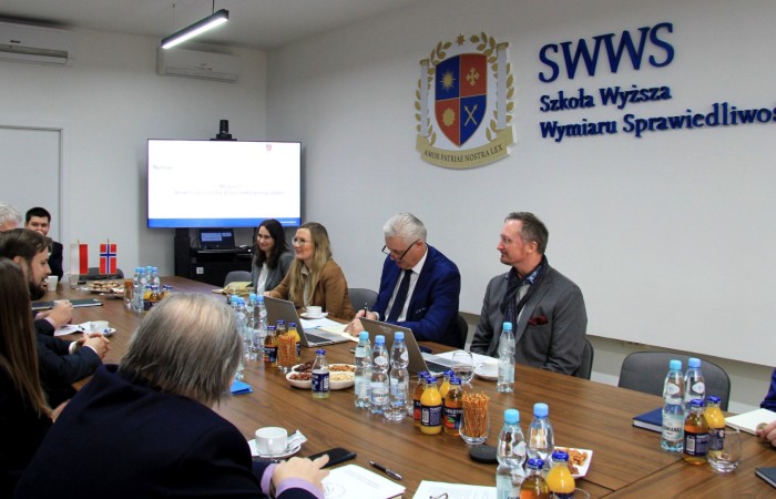 A group of people talking at the big table. On the table, there are laptops, drinks and documents. In the background, there's a TV and The Academy of Justice's coat of arms. A group of people talking at the big table. On the table, there are laptops, drinks and documents. In the background, there's a TV and The Academy of Justice's coat of arms.