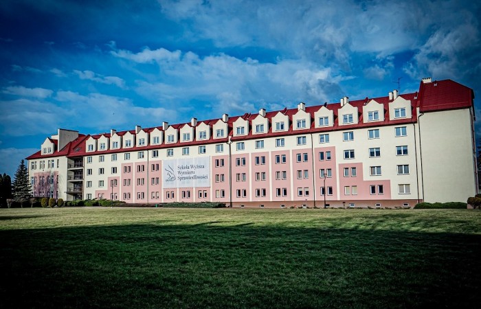 Na zdjęciu widoczny duży budynek domu akademickiego z wieloma oknami. Na środku budynku widnieje baner z nazwą uczelni i jej godłem. Na zdjęciu widoczny duży budynek domu akademickiego z wieloma oknami. Na środku budynku widnieje baner z nazwą uczelni i jej godłem.