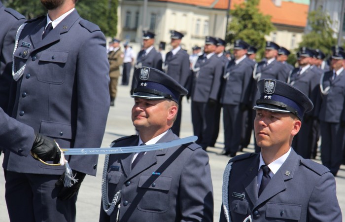 Grupa funkcjonariuszy polskiej Służby Więziennej bierze udział w oficjalnej uroczystości na placu. Dwoje funkcjonariuszy klęczy, pozostali w pozycji 