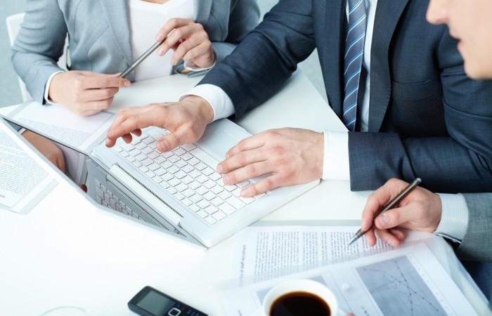 Grupa trzech ludzi w garniturach pracuje z laptopem przy stole. Na blacie stołu dokumenty i telefon komórkowy.