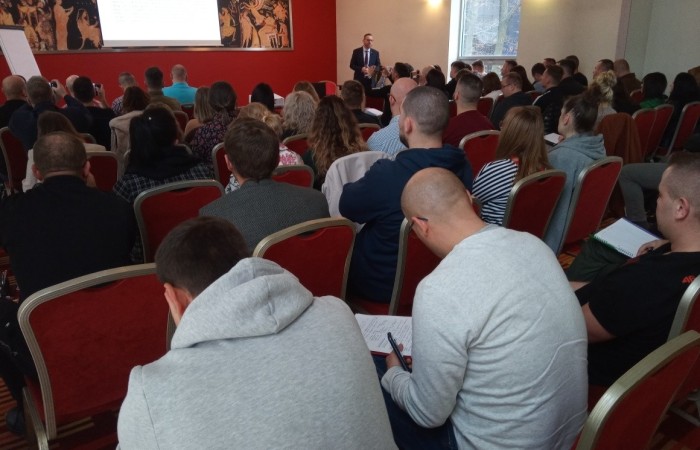 Duża grupa słuchaczy siedzi na krzesłach i przysłuchuje się prezentacji prowadzącego ubranego w strój formalny. Duża grupa słuchaczy siedzi na krzesłach i przysłuchuje się prezentacji prowadzącego ubranego w strój formalny.