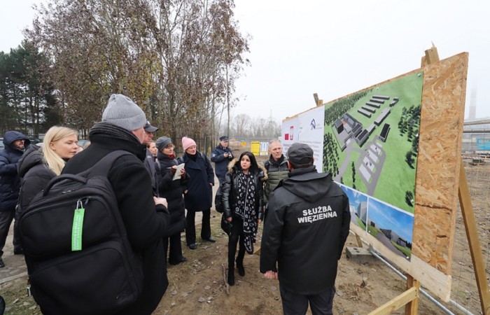 Funkcjonariusz Służby Więziennej oprowadza grupę osób po terenie budowy. Po prawej stronie plakat z wizualizacją inwestycji. Funkcjonariusz Służby Więziennej oprowadza grupę osób po terenie budowy. Po prawej stronie plakat z wizualizacją inwestycji.