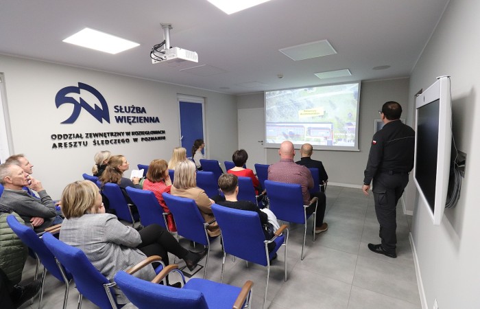 Grupa osób siedząca na krzesłach w budynku Służby Więziennej ogląda prezentację wyświetloną za pomocą rzutnika. Po prawej stronie funkcjonariusz SW Grupa osób siedząca na krzesłach w budynku Służby Więziennej ogląda prezentację wyświetloną za pomocą rzutnika. Po prawej stronie funkcjonariusz SW