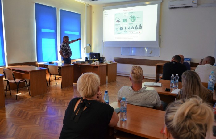 Grupa osób zajmuje miejsca po prawej stronie sali wykładowej. Na ich biurkach notatniki oraz butelki wody. Po lewej stronie sali prowadzący wskazuje palcem na prezentację multimedialną. Grupa osób zajmuje miejsca po prawej stronie sali wykładowej. Na ich biurkach notatniki oraz butelki wody. Po lewej stronie sali prowadzący wskazuje palcem na prezentację multimedialną.