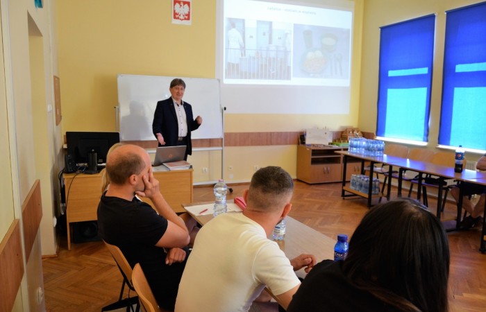 Na pierwszym planie grupa osób przysłuchuje się prezentacji prowadzącego zajęcia. W tle wyświetlona prezentacja multimedialna na ścianie sali. Na pierwszym planie grupa osób przysłuchuje się prezentacji prowadzącego zajęcia. W tle wyświetlona prezentacja multimedialna na ścianie sali.